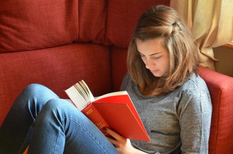 Teen in gray shirt and blue jeans reading a book with a red cover on a red seat